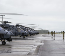 Nh90 helicopters line up on the flight line in cloudy weather. The ground is also wet. Aviators in uniform and helmets stand with thumbs up and arms out towards the aircraft.