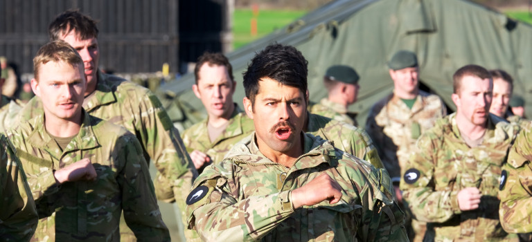 Soldiers in camouflage uniform with a black patch on their right arm with a white kiwi on it do the haka.