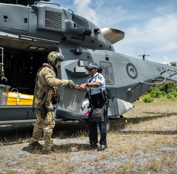 A uniformed personnel hands over a to a police officer standing outside an NH90 helicopter. 