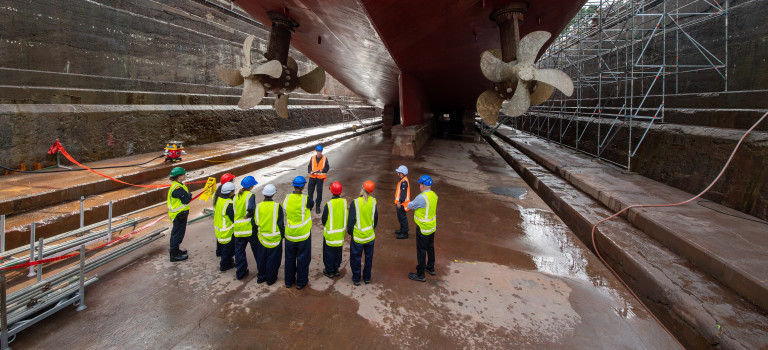 Students in high vis jackets and hard hard hats stand in a drydock looking up the stern of a large ship that has two large propellors.