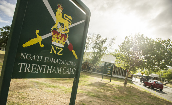 A sign with the New Zealand Army logo and Ngāti Tūmatauenga Trentham Camp written on it. The sun is shining and trees and the entrance to the camp and in the background. 