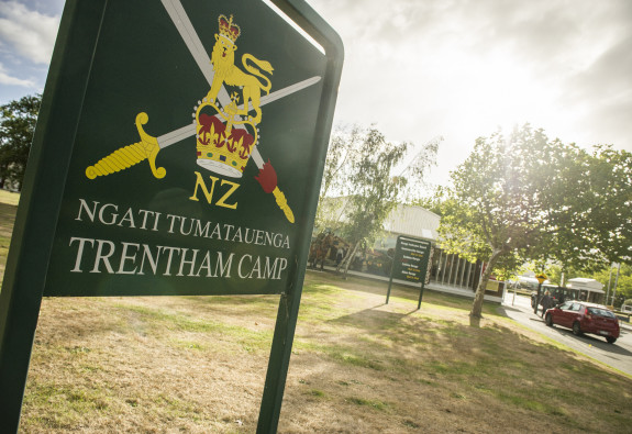 A sign with the New Zealand Army logo and Ngāti Tūmatauenga Trentham Camp written on it. The sun is shining and trees and the entrance to the camp and in the background. 