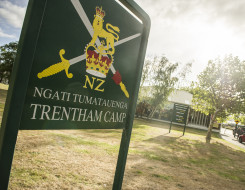 A sign with the New Zealand Army logo and Ngāti Tūmatauenga Trentham Camp written on it. The sun is shining and trees and the entrance to the camp and in the background. 