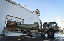 Royal New Zealand Navy's HMNSZ Canterbury berthed as a New Zealand Army MHOV drives off the ship. In the background there is blue sky and a crane as part of the port in Wellington. 