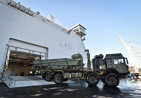 Royal New Zealand Navy's HMNSZ Canterbury berthed as a New Zealand Army MHOV drives off the ship. In the background there is blue sky and a crane as part of the port in Wellington. 