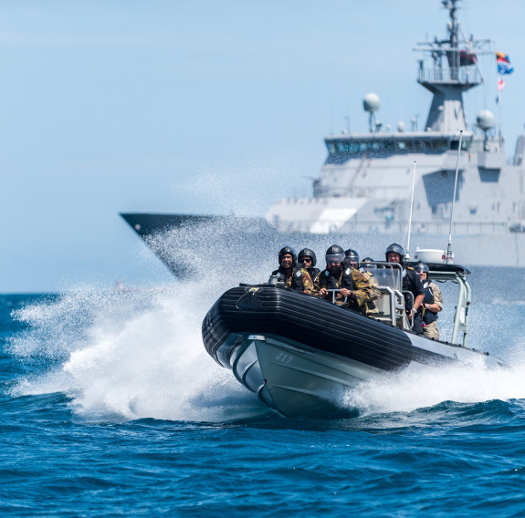 A Royal New Zealand Navy Rigid Hull Inflatable Boat moves through the water with uniformed people on board. HMNZS Wellington is in the background.