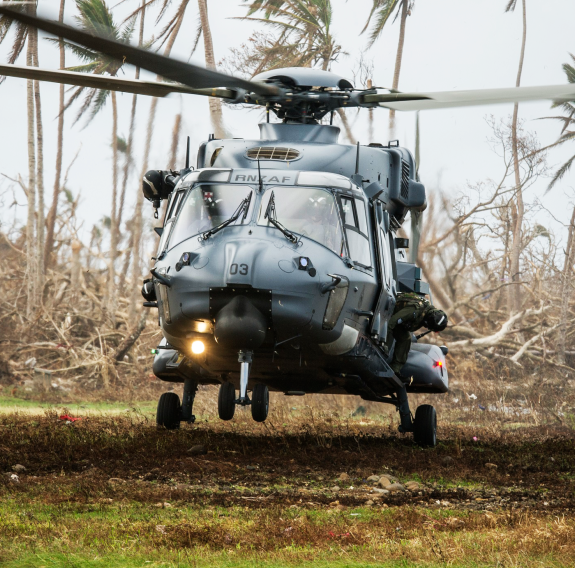 A Royal New Zealand Air Force NH90 helicopter lands on the grass in Fiji, palm trees blowing the background as well as mess from Cyclone Winston