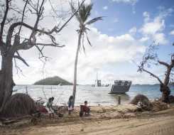 A Royal New Zealand Navy Landing Craft Mehchniciam (LCM) in the ocean makes its way to the shore. In the background you see an island with some trees on it, clouds and some blue sky. In the foreground some children sit with a wheelbarrow and wait for the 