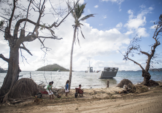 A Royal New Zealand Navy Landing Craft Mehchniciam (LCM) in the ocean makes its way to the shore. In the background you see an island with some trees on it, clouds and some blue sky. In the foreground some children sit with a wheelbarrow and wait for the 