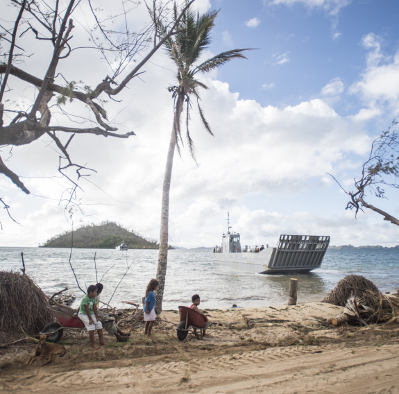 A Royal New Zealand Navy Landing Craft Mehchniciam (LCM) in the ocean makes its way to the shore. In the background you see an island with some trees on it, clouds and some blue sky. In the foreground some children sit with a wheelbarrow and wait for the 