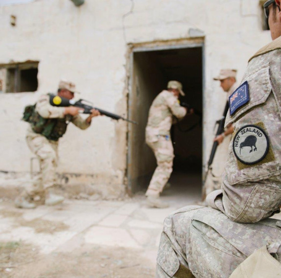 A New Zealand Army soldier sits to the right of the image and watches other four uniformed soldiers enter a building with weapons.