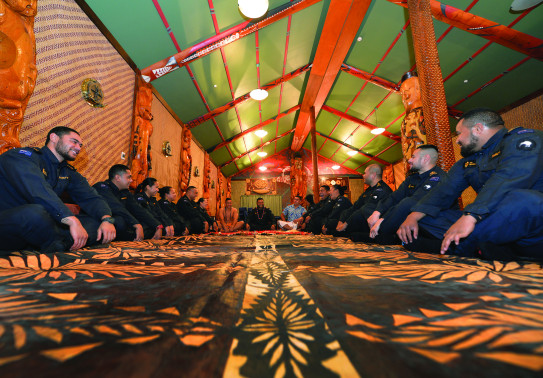 A group of sailors sit on the ground in a semi-cycle of the Royal New Zealand Navy marae with smiles on their faces and some laughing.  