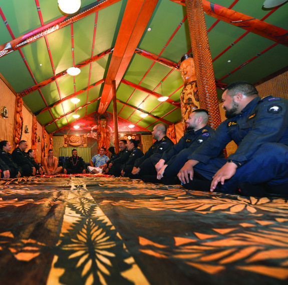 A group of sailors sit on the ground in a semi-cycle of the Royal New Zealand Navy marae with smiles on their faces and some laughing.  