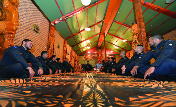 A group of sailors sit on the ground in a semi-cycle of the Royal New Zealand Navy marae with smiles on their faces and some laughing.  