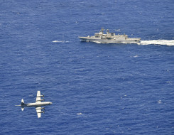 A Royal New Zealand Air Force P-3K2 Orion flies over the blue ocean in the bottom left side of the photo with HMNZS Te Kaha during a firing exercise 