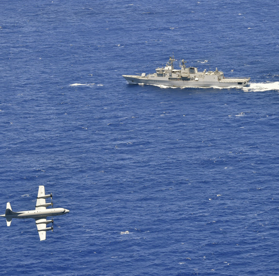 A Royal New Zealand Air Force P-3K2 Orion flies over the blue ocean in the bottom left side of the photo with HMNZS Te Kaha during a firing exercise 