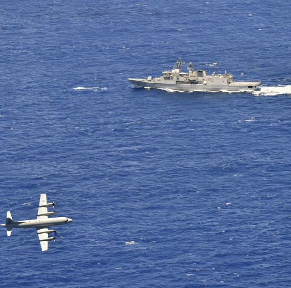 A Royal New Zealand Air Force P-3K2 Orion flies over the blue ocean in the bottom left side of the photo with HMNZS Te Kaha during a firing exercise 