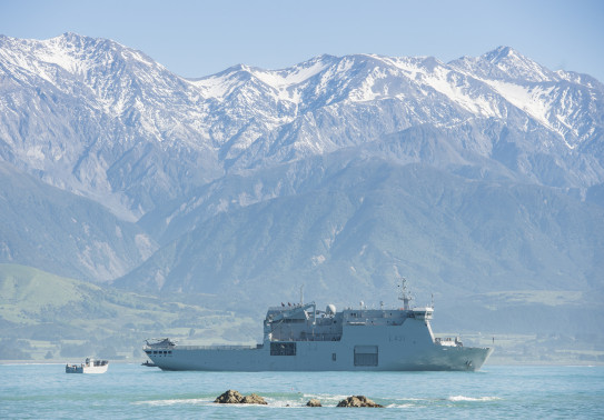HMNZS Canterbury sits in the ocean off the coast of Kaikōura with the Kaikōura ranges in the background covered in snow. It's a sunny day. 