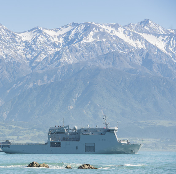 HMNZS Canterbury sits in the ocean off the coast of Kaikōura with the Kaikōura ranges in the background covered in snow. It's a sunny day. 
