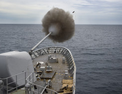 Royal New Zealand Navy's HMNZS Te Kaha fires the 5-inch gun. In the image you see a puff of smoke and the shell. Grey sky. 