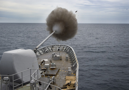 Royal New Zealand Navy's HMNZS Te Kaha fires the 5-inch gun. In the image you see a puff of smoke and the shell. Grey sky. 