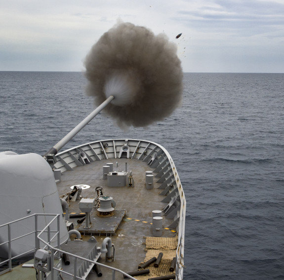 Royal New Zealand Navy's HMNZS Te Kaha fires the 5-inch gun. In the image you see a puff of smoke and the shell. Grey sky. 