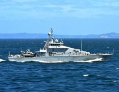 Royal New Zealand Navy's HMNZS Taupō sailing in the ocean on a nice day, blue sky in the background and some hills. 