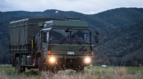 A New Zealand Army Medium and Heavy Operational Vehicle (MHOV) with lights on sits in a grassy area with a green covered hill in the background. It's a darker image with grey sky.