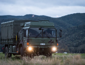 A New Zealand Army Medium and Heavy Operational Vehicle (MHOV) with lights on sits in a grassy area with a green covered hill in the background. It's a darker image with grey sky.