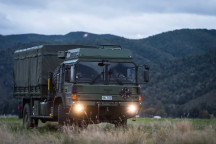 A New Zealand Army Medium and Heavy Operational Vehicle (MHOV) with lights on sits in a grassy area with a green covered hill in the background. It's a darker image with grey sky.