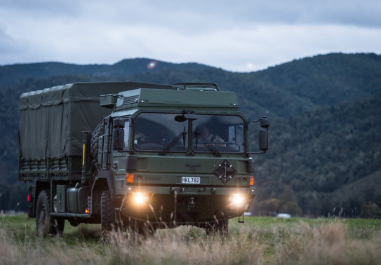 A New Zealand Army Medium and Heavy Operational Vehicle (MHOV) with lights on sits in a grassy area with a green covered hill in the background. It's a darker image with grey sky.