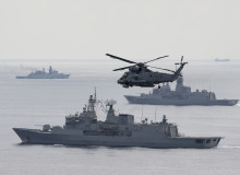 A SH-2G(I) Seasprite Helicopter flies in the foreground of the image with HMNZS Te Kaha. In the background there are two other warships from other countries