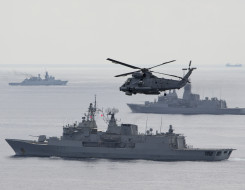 A SH-2G(I) Seasprite Helicopter flies in the foreground of the image with HMNZS Te Kaha. In the background there are two other warships from other countries