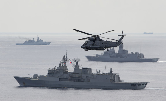 A SH-2G(I) Seasprite Helicopter flies in the foreground of the image with HMNZS Te Kaha. In the background there are two other warships from other countries