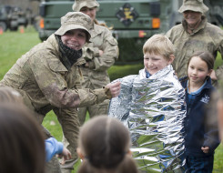 A New Zealand Army soldier engages with children at a school visit. The child is being wrapped in am emergency blanket as they demonstrate how to use it. In the background there are other soldiers and Army capability. In the foreground, heads of other stu