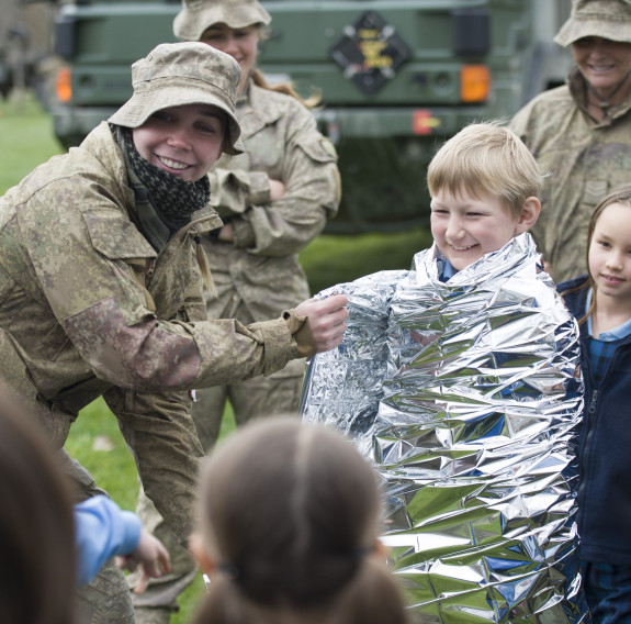 A New Zealand Army soldier engages with children at a school visit. The child is being wrapped in am emergency blanket as they demonstrate how to use it. In the background there are other soldiers and Army capability. In the foreground, heads of other stu
