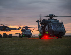 Three Royal New Zealand Air Force NH90 helicopters at dusk. One aircraft in the background is in flight, the other two are on the ground. The sky is an orange morning sunrise.