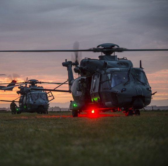 Three Royal New Zealand Air Force NH90 helicopters at dusk. One aircraft in the background is in flight, the other two are on the ground. The sky is an orange morning sunrise.