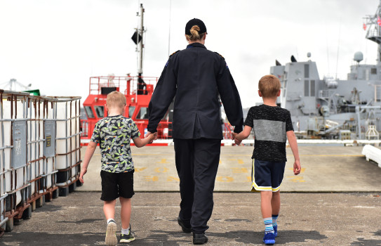 The back of a sailor that is walking away from the camera holding the hands of two children with each of their hands. Walking towards a ship on the docks at Devonport Naval Base. 