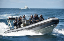 A Royal New Zealand Navy Rigid-Hulled Inflatable Boat (RHIB) full with sailors wearing helmets moves through the ocean with a ship in the background on a nice day, blue sky.