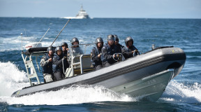 A Royal New Zealand Navy Rigid-Hulled Inflatable Boat (RHIB) full with sailors wearing helmets moves through the ocean with a ship in the background on a nice day, blue sky.