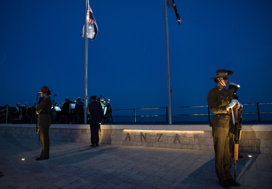 NZDf personnel stand in a catafalque guard at the Anzac Commemorative Site in Gallipoli. Thee New Zealand and Australian flags are flying from flag poles and it is dark as the ceremony is at dawn.