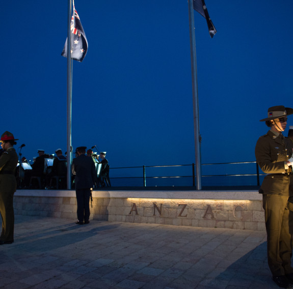 NZDf personnel stand in a catafalque guard at the Anzac Commemorative Site in Gallipoli. Thee New Zealand and Australian flags are flying from flag poles and it is dark as the ceremony is at dawn.