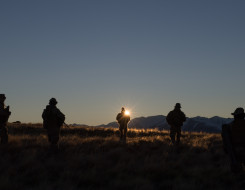 Five New Zealand Army soldiers on patrol on a hill. The sun is rising in the background (right behind the soldier in the middle). The soldiers are silhouetted and you can see the hills in the backgroun. 