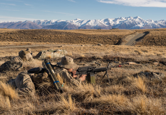 New Zealand Army soldiers lie in the tussock as they train in the Tekapo Military Training Area. You can see the snow capped mountains in the background, you can also see the road.