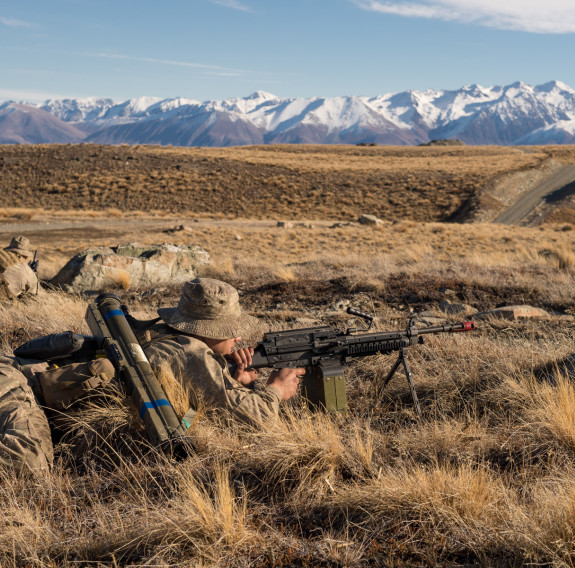 New Zealand Army soldiers lie in the tussock as they train in the Tekapo Military Training Area. You can see the snow capped mountains in the background, you can also see the road.