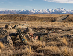 New Zealand Army soldiers lie in the tussock as they train in the Tekapo Military Training Area. You can see the snow capped mountains in the background, you can also see the road.