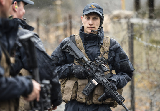 A Royal New Zealand Air Force airman carrying a MARS-L weapon wearing a headset and Air Force cap. To the left in the foreground two other airmen look away from the camera and are out of focus. They are wet from the rain. In the background you can see a b