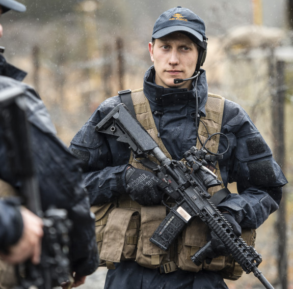 A Royal New Zealand Air Force airman carrying a MARS-L weapon wearing a headset and Air Force cap. To the left in the foreground two other airmen look away from the camera and are out of focus. They are wet from the rain. In the background you can see a b