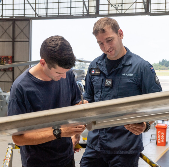 Two Royal New Zealand Air Force avionic technicians look at a piece of equipment in an aircraft hanger. There is a helicopter in the background and a door opening to outside. 